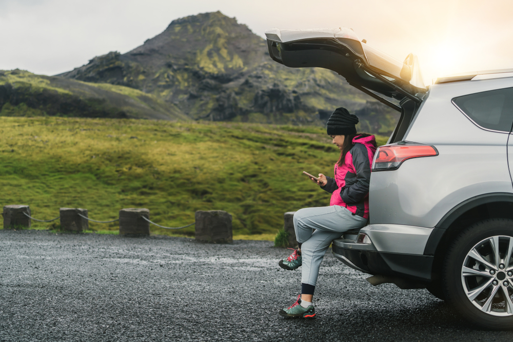 Blue Car Rental full cover service. Woman sitting on the trunk of a rental car in Iceland waiting for assistance.